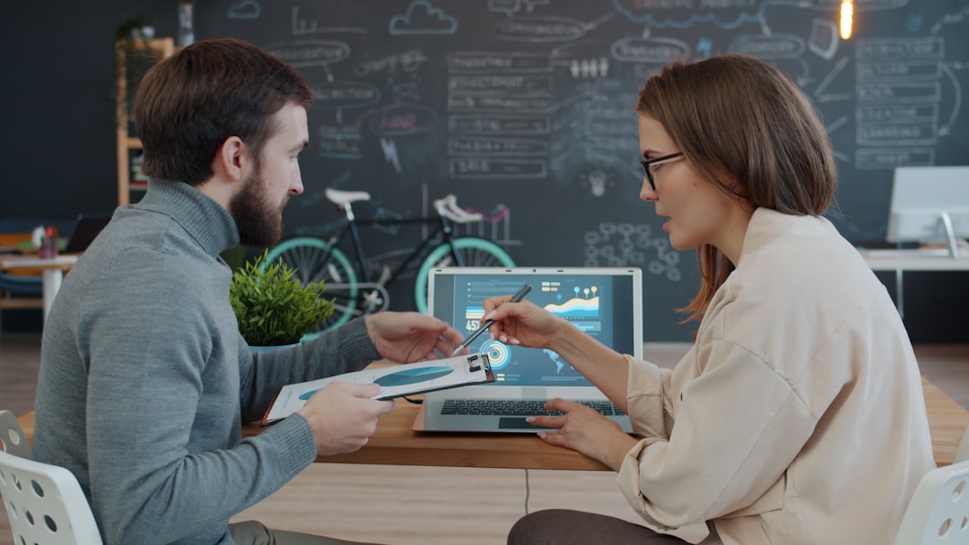 Man and woman colleagues are discussing business looking at laptop screen in modern office sitting at desk together. People, communication and gadgets concept.